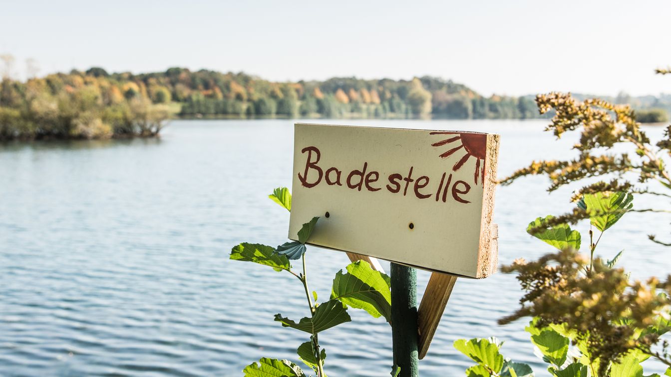 Das Bild zeigt ein Schlid mit der Aufschrift "Badestelle" am Ufer des Albrechtshainer See. Im Hintergrund sieht man das Wasser und den nahegelegenen Wald.