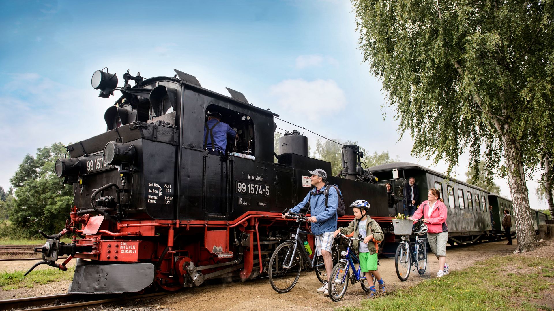 Eine Familie steht mit ihren Fahrrädern an einem sommerlichen Tag vor einer Dampfeisenbahn in der Leipzig Region.